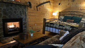 Interior acogedor de una cabaña con chimenea de piedra, una cama con cabecera temática de bosque y una pequeña mesa con una planta en maceta.