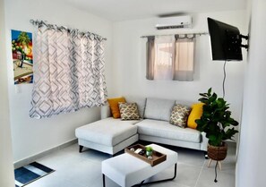 A modern living room with a sectional sofa, a small white ottoman, a potted plant, and a mounted TV.