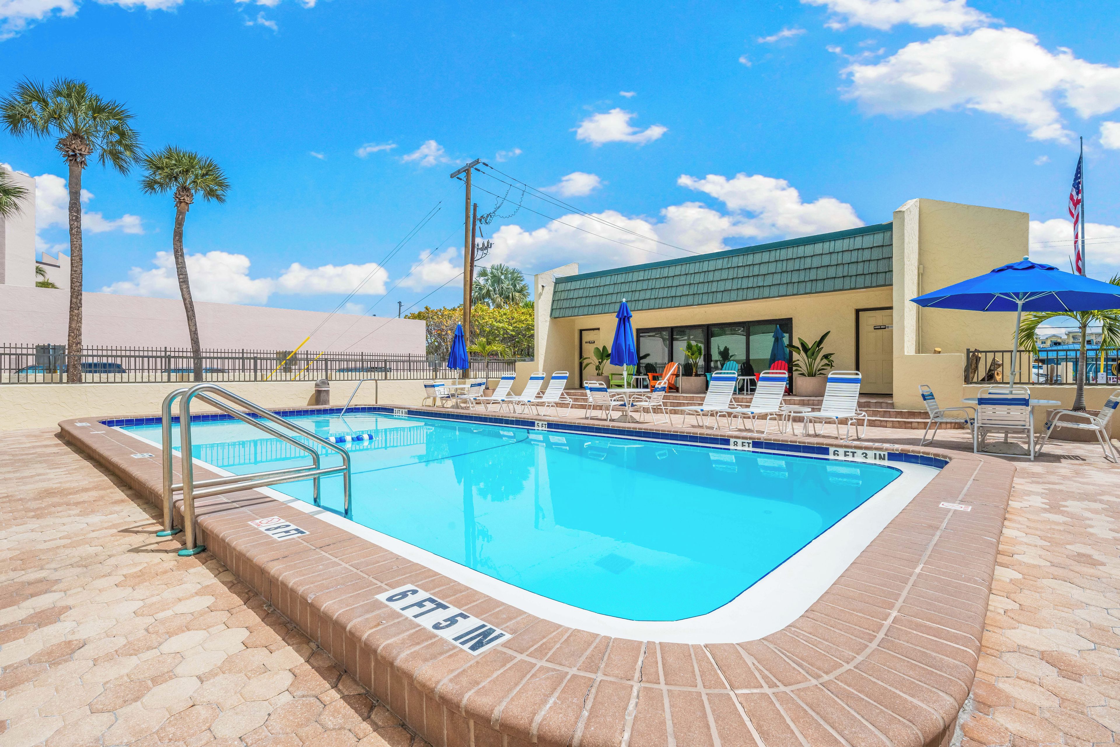 A rectangular swimming pool with a brick edge, surrounded by lounge chairs and umbrellas, under a clear sky.