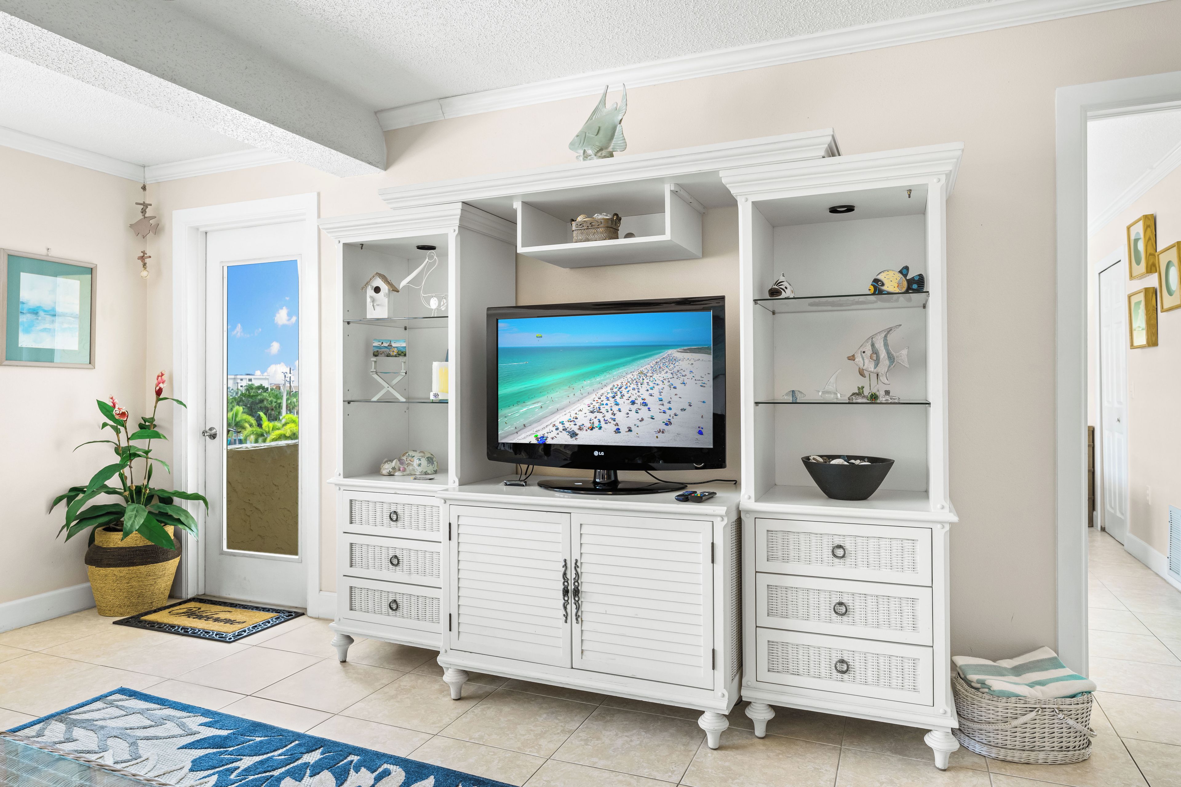 A living room with a television displaying a beach scene, white cabinetry with multiple drawers, a wicker basket, and a potted plant.