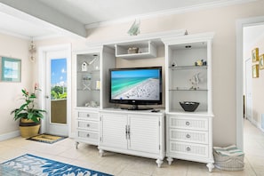 A living room with a television displaying a beach scene, white cabinetry with multiple drawers, a wicker basket, and a potted plant. - Sea Shell by Beachside Management (Siesta Key)