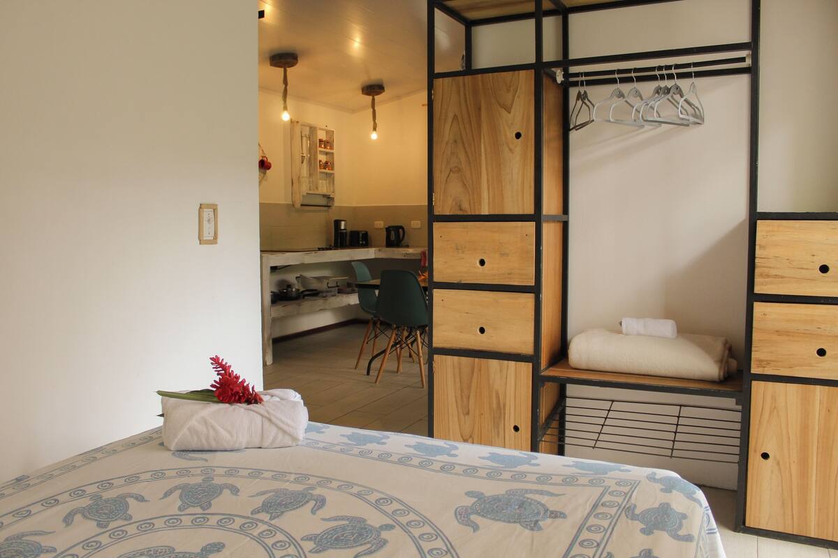 Un dormitorio con una cama, un armario de madera y una kitchenette al fondo.