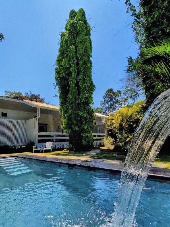 A swimming pool with a waterfall feature, surrounded by greenery and a building with a balcony.