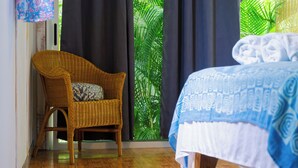 Une pièce avec un fauteuil en osier, un lit recouvert d’une courtepointe à motifs et une vue sur un feuillage verdoyant luxuriant, visible à travers de grandes fenêtres.