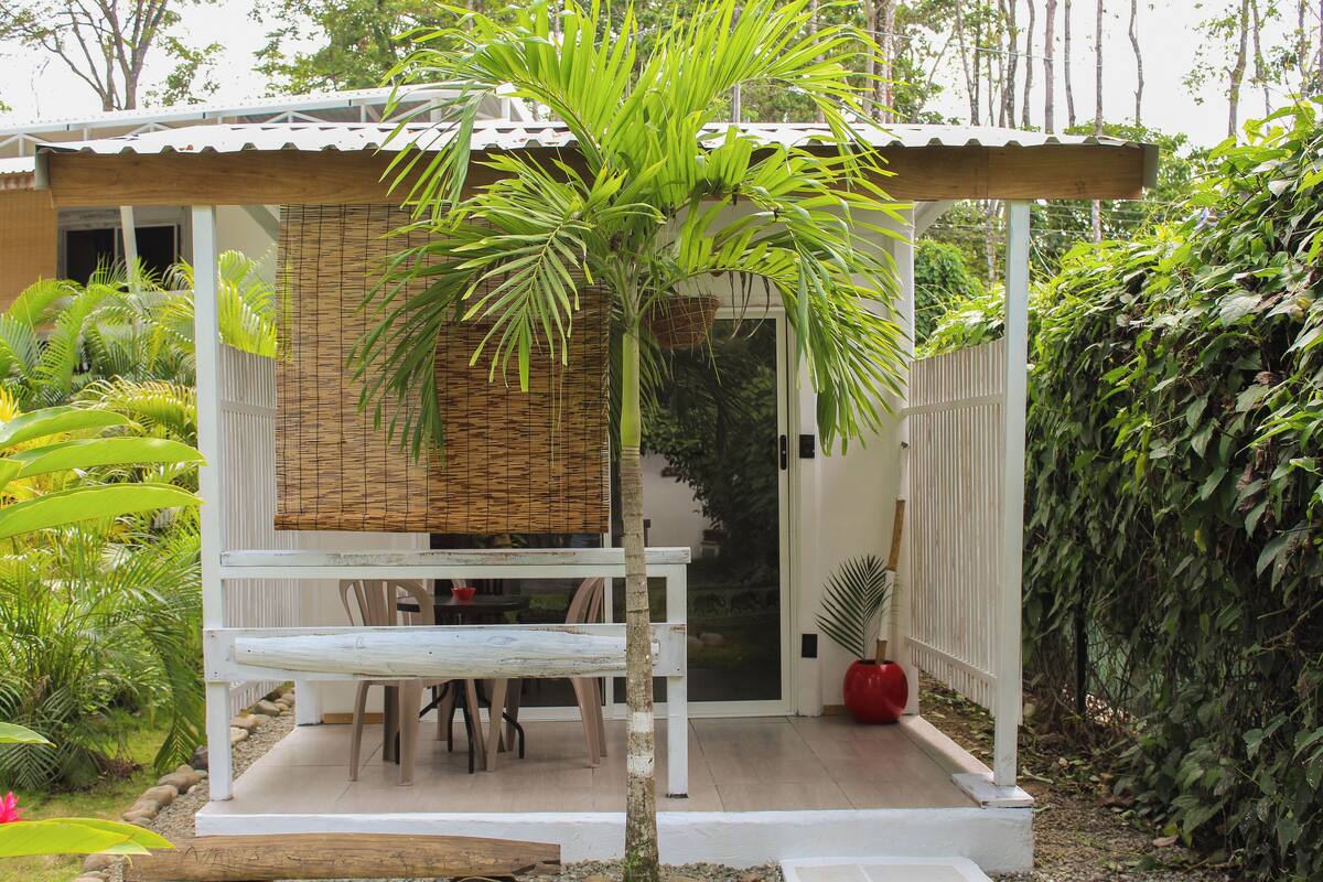 Un espacio de estar al aire libre techado con una mesa y sillas, rodeado de abundante vegetación y un pequeño árbol.