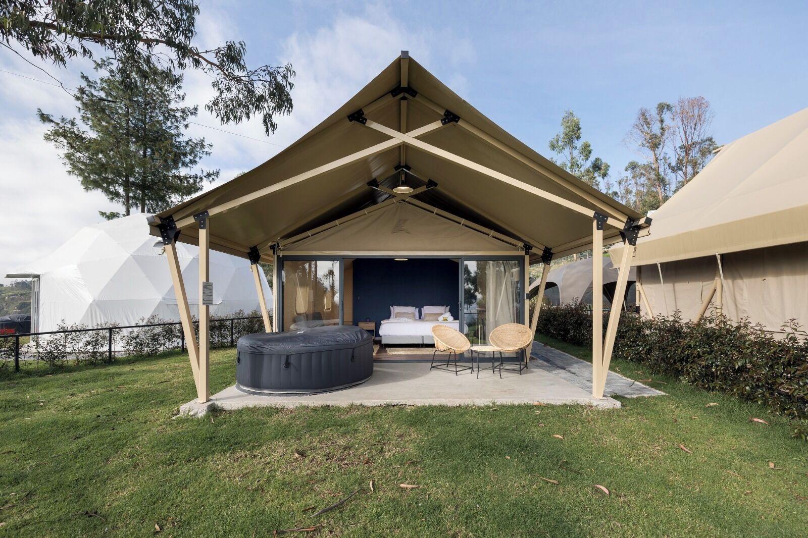 A tented outdoor accommodation with a bed, a hot tub, and two wicker chairs.
