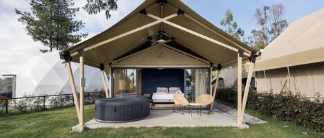 A tented outdoor accommodation with a bed, a hot tub, and two wicker chairs.