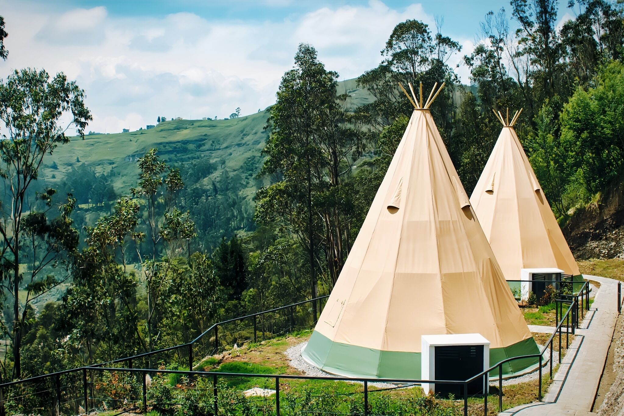 Deux tipis beiges à la base verte, entourés d’une végétation luxuriante et de collines.
