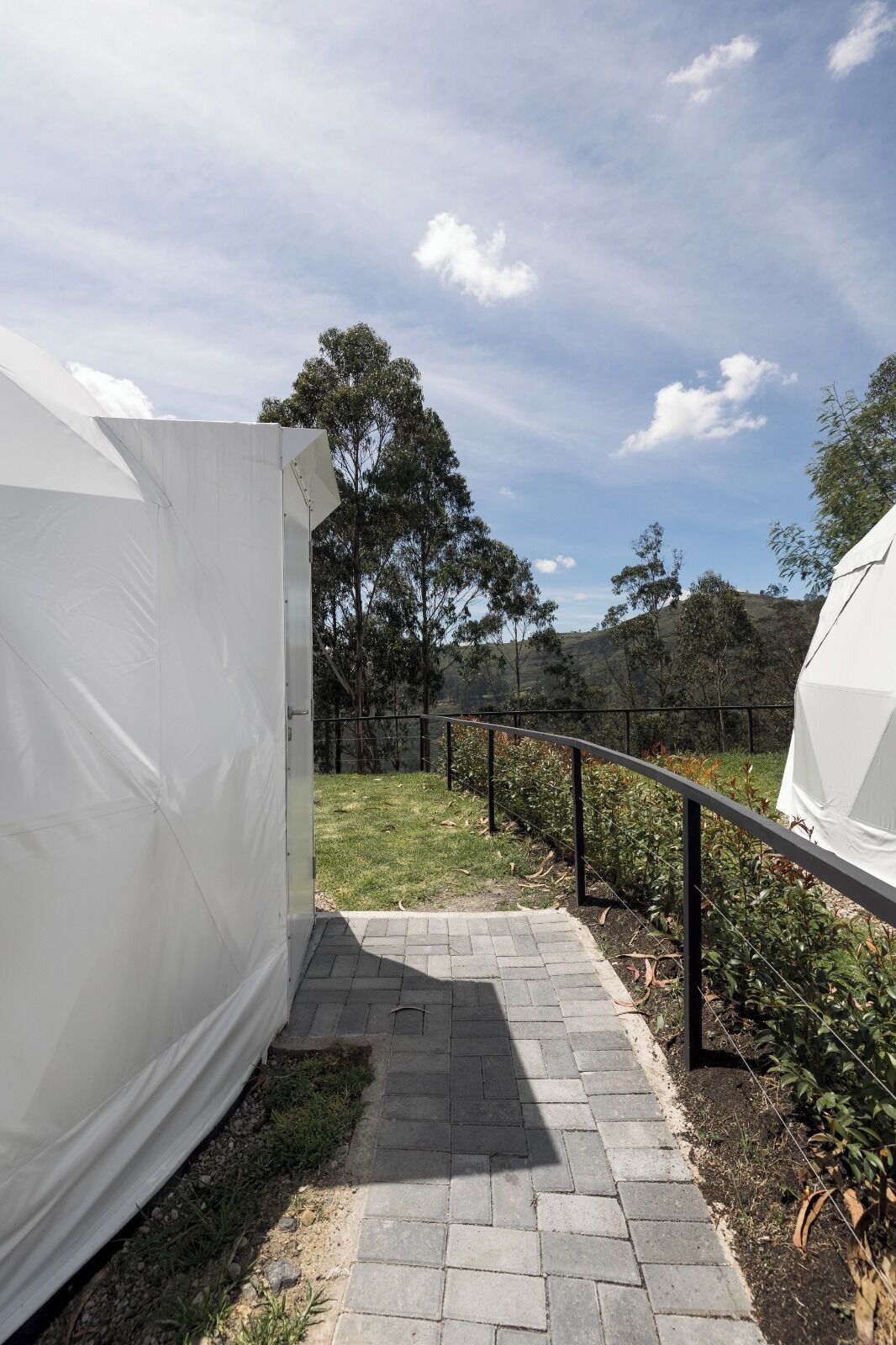 A pathway leading to a white tent-like structure with a metal railing, surrounded by greenery and trees.
