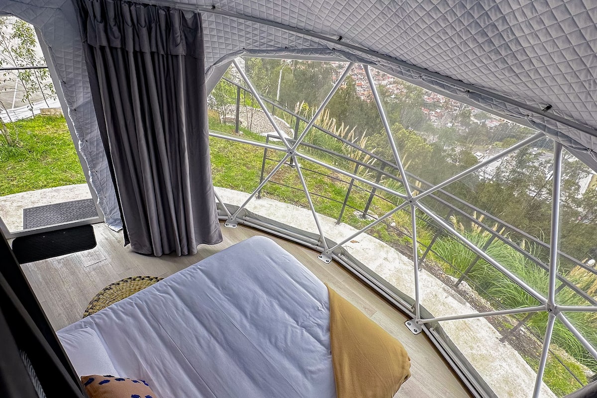 A modern, spacious room with a large bed, a view of greenery and buildings, and a unique geometric ceiling design.