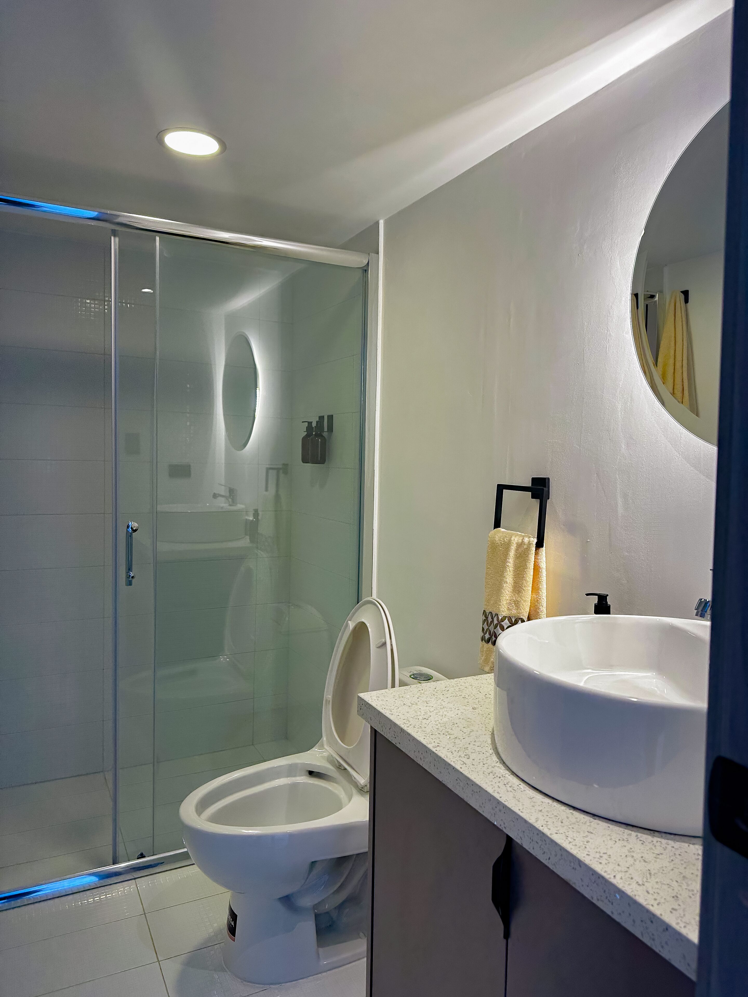 Un baño moderno con cabina de ducha de vidrio, inodoro blanco y un lavamanos grande y redondo con espejo superior.
