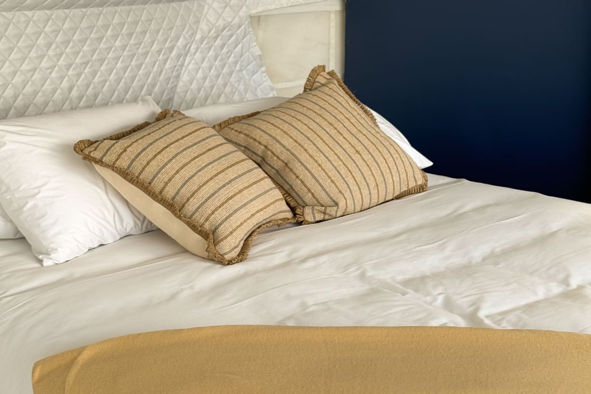 A bed with white bedding, two striped pillows, and a beige throw blanket.