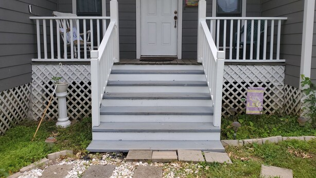 A gray house with a white front door, two windows, a porch with white railings, and a small garden area.