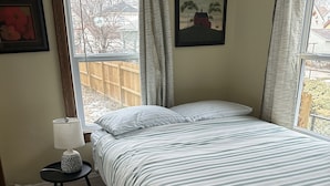 A bedroom with a bed, a nightstand, a window with curtains, and a wooden floor. - Stella's Sweet Place (Minneapolis)