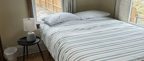 Een slaapkamer met een bed, een nachtkastje, een raam met gordijnen en een houten vloer.