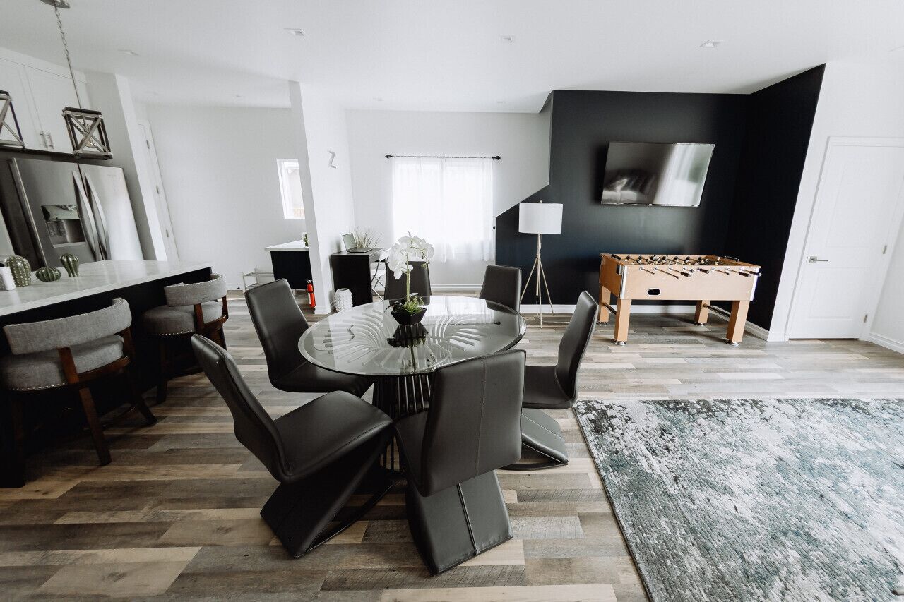 Un salon moderne avec une table ronde en verre, des fauteuils en cuir noir, un baby-foot et un téléviseur à écran plat fixé sur un mur noir.