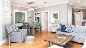 A spacious living room with a wooden coffee table, grey sofas, and a dining area with a wooden table and chairs. - Cute Cottage~2 Pools~Beach Nearby ~ Book Now for a Relaxing Stay! (Gulf Shores)