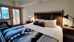 A bedroom with a large bed, a wooden headboard, a window with blinds, a desk with a lamp, and a digital clock.
