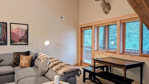 A cozy living room with a gray sectional sofa, a round coffee table, and a wooden bench. A deer head is mounted on the wall, and there are framed posters on the wall.