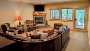 Un salón acogedor con chimenea de piedra, sofás de cuero, televisor de pantalla plana y ventanas grandes con vistas al exterior.