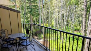 A wooden deck with a table and chairs, surrounded by a metal railing, overlooking a forested area.