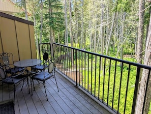 A wooden deck with a table and chairs, surrounded by a metal railing, overlooking a forested area.