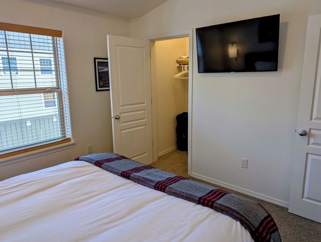 A bedroom with a bed, a TV mounted on the wall, a closet, and a window with blinds.