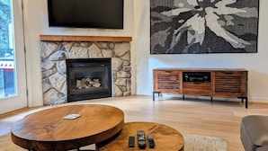 A living room with a stone fireplace, a flat-screen TV mounted above it, a large abstract painting, and a wooden entertainment unit. - Chalet 73 at the Official Collins Lake Resort (Government Camp)