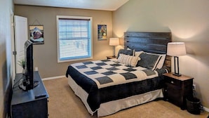 A bedroom with a checkered bedspread, a ceiling fan, a window with blinds, and a nightstand with a lamp.
