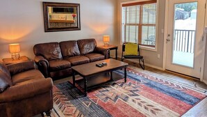A living room with a brown leather sofa, a coffee table, a patterned rug, a side table with a lamp, and a window with blinds. - Chalet 133 at the Official Collins Lake Resort (Government Camp)