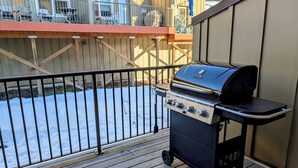 Un barbecue sur une terrasse en bois, avec vue sur un bâtiment et une zone délimitée par une clôture.