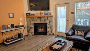 A living room with a stone fireplace, a television mounted above it, a leather sofa, and a coffee table with a tray. - Chalet 35 at the Official Collins Lake Resort (Government Camp)