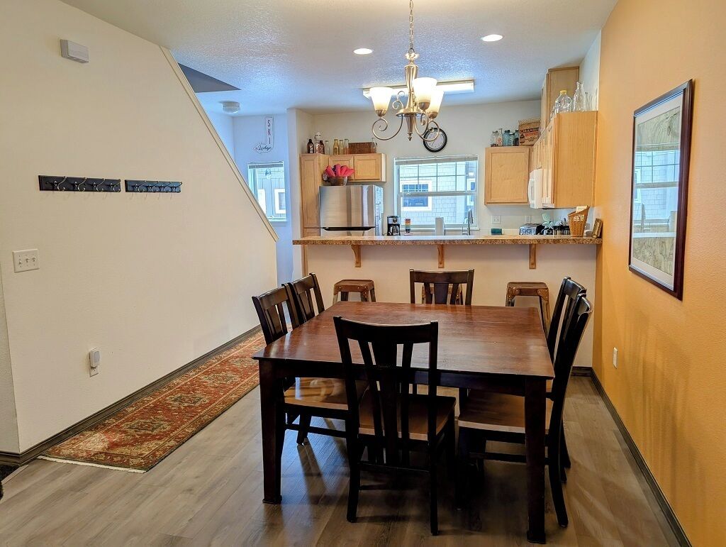 A dining area with a wooden table and chairs, a chandelier, and a kitchen area with wooden cabinets and a refrigerator.