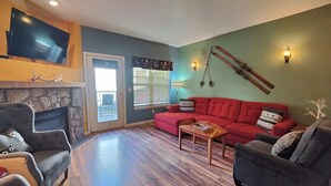 A living room with a stone fireplace, a red sofa, and a television mounted on the wall. - Chalet 68 at the Official Collins Lake Resort (Government Camp)