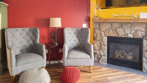 Two grey tufted armchairs, a red knitted ottoman, a stone fireplace, and a yellow wall with a TV. - Chalet 68 at the Official Collins Lake Resort (Government Camp)