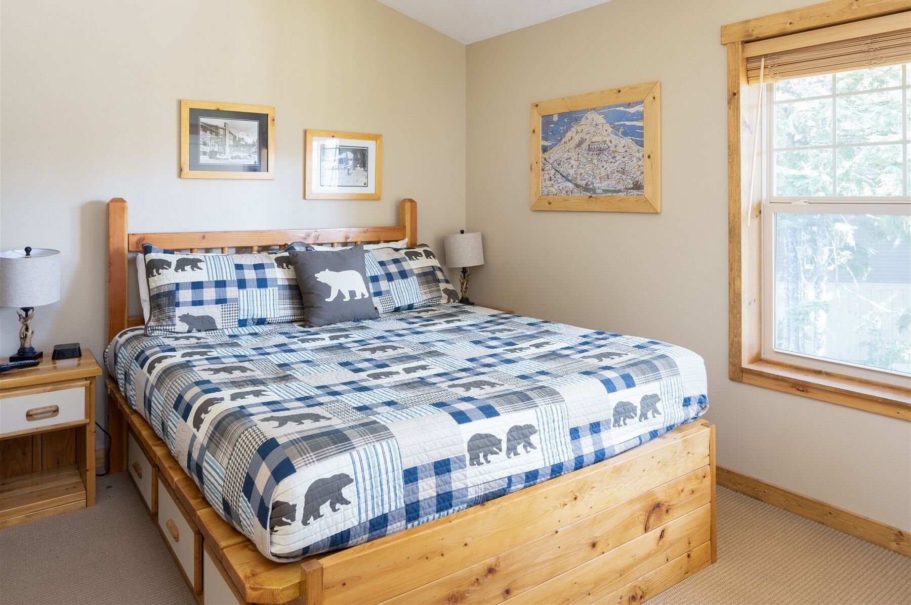 Un dormitorio con estructura de cama de madera, colcha cuadriculada con motivos de osos, mesita de noche con lámpara y ventana con vista a árboles.
