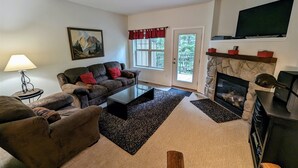 A living room with a sofa, armchair, fireplace, television, and a painting on the wall. - Chalet 103 at the Official Collins Lake Resort (Government Camp)