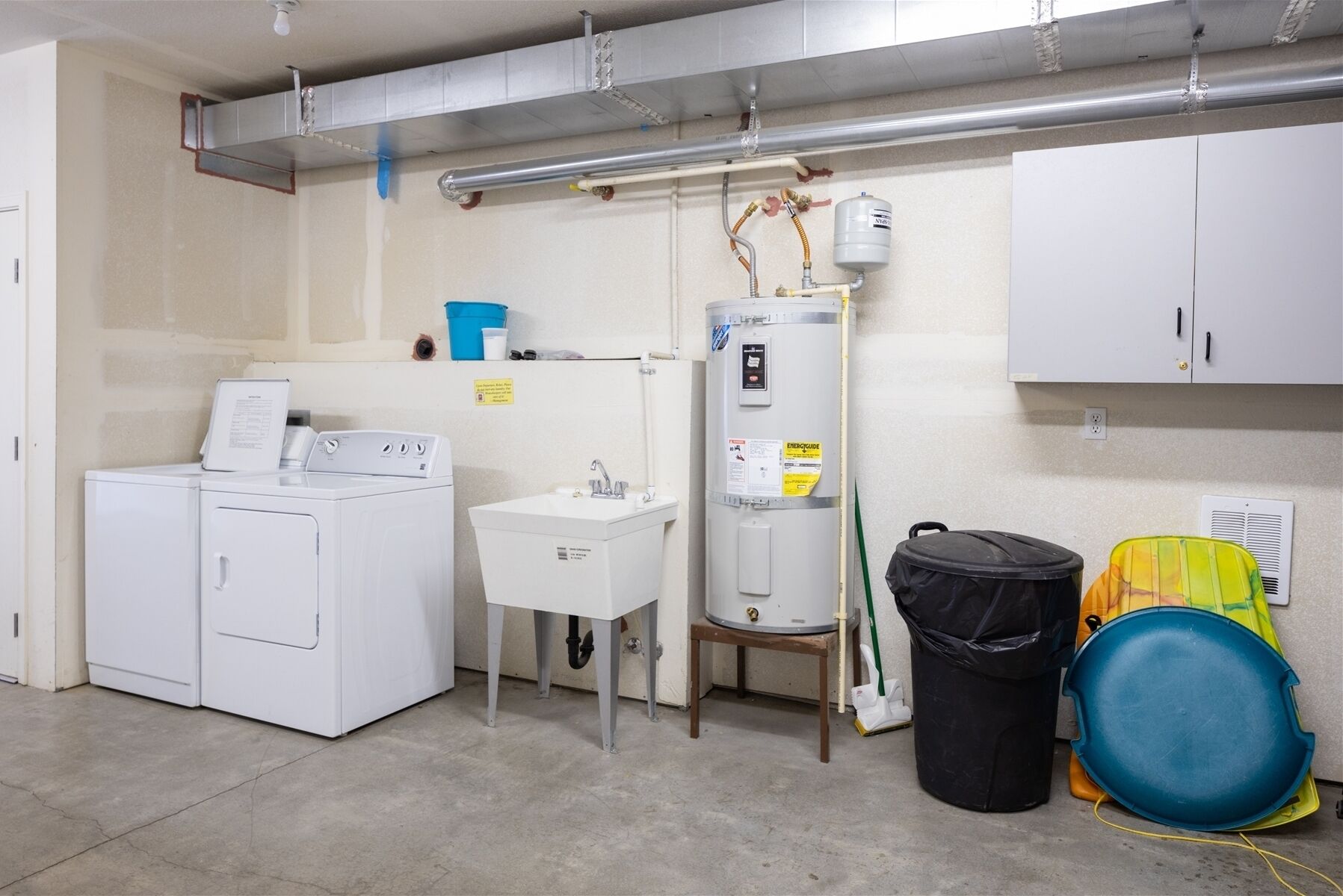 Ein Waschraum mit Waschmaschine und Trockner, einem Waschbecken, einem Warmwasserbereiter und einem Mülleimer.