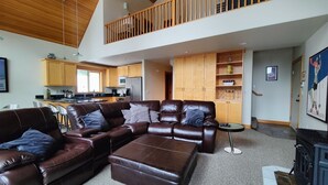 A spacious living room with a large brown leather sectional sofa, a small round coffee table, and a fireplace. There is a kitchen in the background with wooden cabinets and a dining area with bar stools. - Slope Side Cabin at the Official Collins Lake Resort (Government Camp)