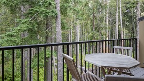 A balcony with a table and two chairs, surrounded by a railing, overlooking a dense forest.