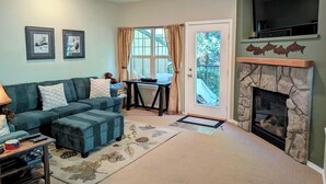 A living room with a sofa, ottoman, fireplace, and a television. - Chalet 106 at the Official Collins Lake Resort (Government Camp)
