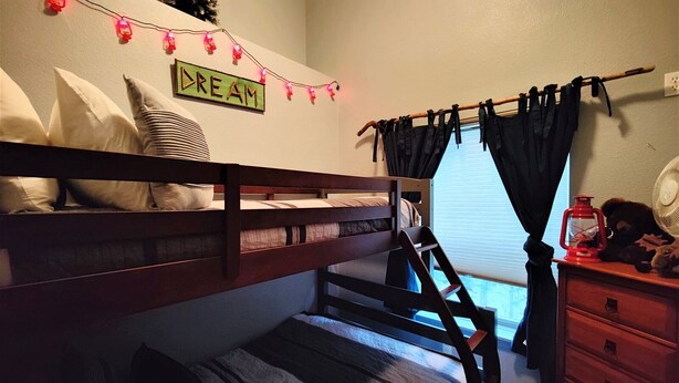 Una cama litera con escalera, un aparador, una mesita con un farol y una ventana con cortinas.