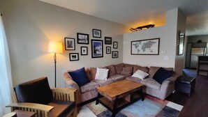 Uma sala de estar com sofá, mesa de centro e luminária. Há quadros nas paredes e um mapa-múndi.
