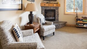 Una sala de estar acogedora con chimenea, dos sillones estampados, una mesita de noche con lámpara y una estantería llena de libros.