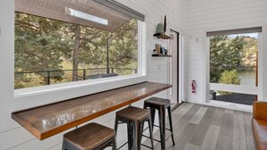 A modern kitchen with a long wooden countertop, two stools, and large windows offering views of trees and a lake.