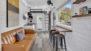 A compact kitchen with a dining area, a loft bedroom, and a large window overlooking a natural landscape.