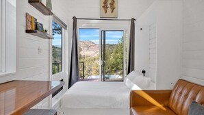 A bedroom with a bed, a wooden desk, a brown leather sofa, and a large window with a mountain view. - Lake Simtustus Resort Juniper Hill Tiny Home #11 - Hot Tub (Madras)