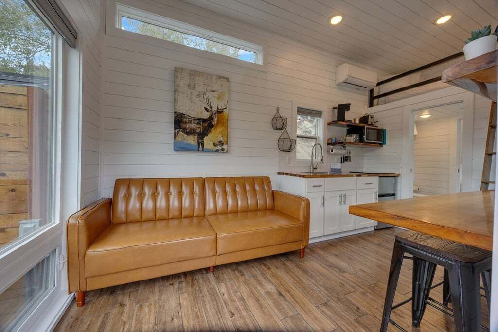A modern kitchen with a wooden floor, white cabinets, a brown leather sofa, and a dining area with a wooden table.