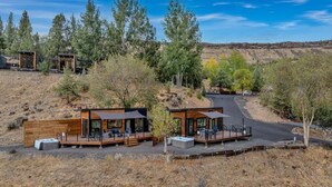 A modern cabin with a deck and outdoor seating area, surrounded by trees and a dry landscape.