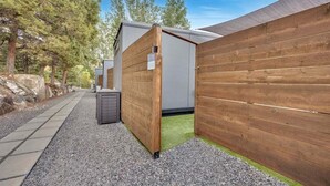Un piccolo spazio esterno recintato da una staccionata in legno, con un vialetto in ghiaia e un edificio dalla facciata grigia.